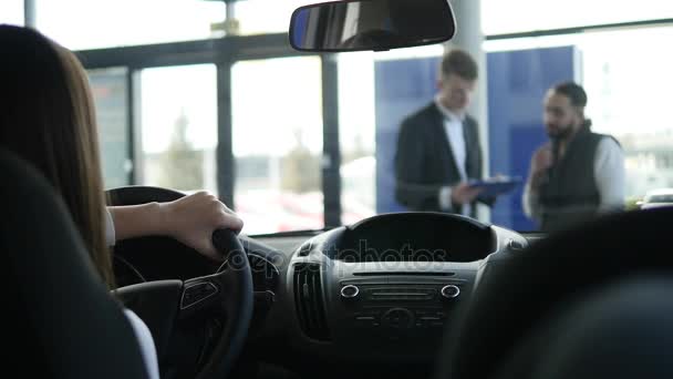 Arrière de la caméra de belle petite fille regardant loin et souriant tout en étant assis dans une nouvelle voiture dans un salon de l'automobile — Video