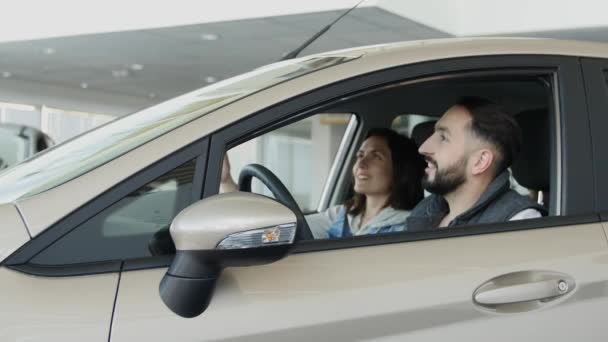 Visite concessionnaire automobile. Belle famille parle et sourit tout en étant assis dans leur nouvelle voiture. jeune homme montre doigt vers le haut — Video