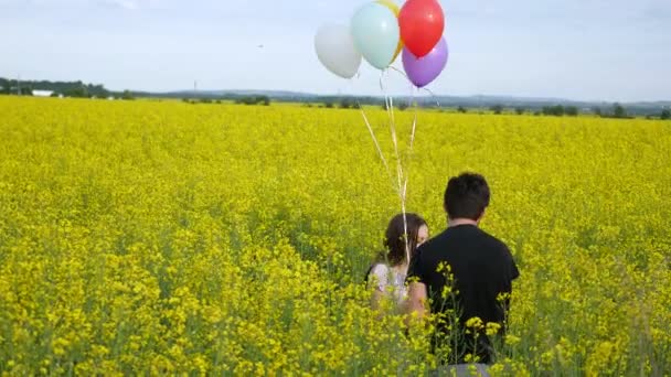 Kleines Mädchen in einem Kleid, das mit Luftballons in der Hand durch ein gelbes Weizenfeld läuft. Zeitlupe — Stockvideo