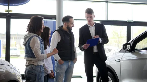 Vendedor de carros mostra uma jovem família informações sobre seu novo carro — Fotografia de Stock