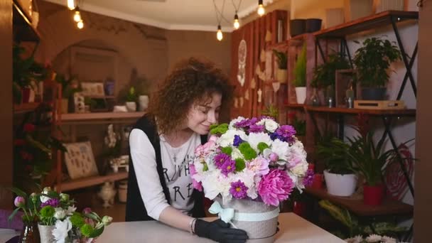 Молода жінка працює флористом у квітковому магазині і дивиться на камеру, посміхається з букетом на столі — стокове відео