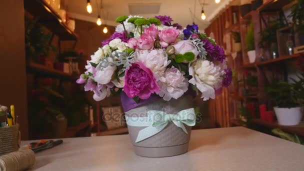 Close-up of bouquet of colorful peonies and roses and blue ribbon on white table. In background flowers shop — Stock Video