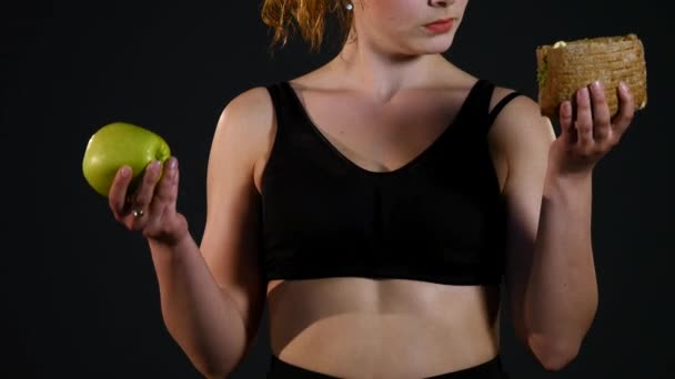 Retrato de una mujer gorda feliz eligiendo entre hamburguesa o manzana aislada sobre un fondo negro — Vídeo de stock