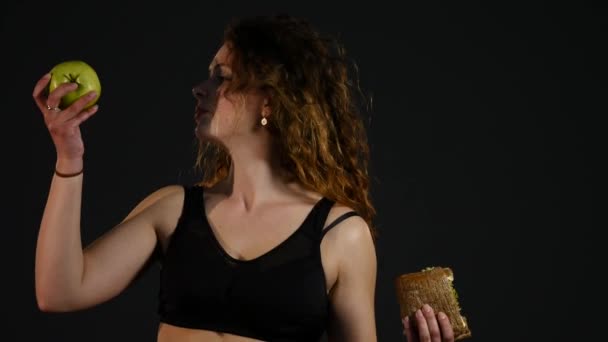 Retrato de una mujer gorda feliz eligiendo entre hamburguesa o manzana aislada sobre un fondo negro — Vídeos de Stock