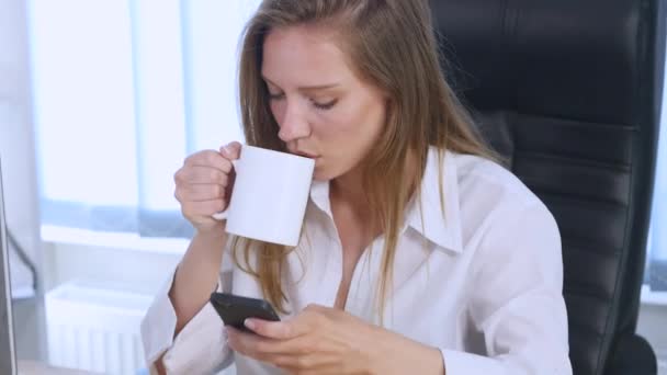 オフィスで笑みを浮かべてコーヒー飲んでいる携帯電話でアプリを使ってスマート フォン女の子。携帯電話での美しい多文化を若いカジュアルな女性プロ — ストック動画