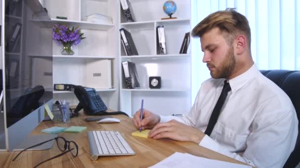 Νέοι επαγγελματίες άνθρωπος εργάζεται στον υπολογιστή και καθιστώντας σημειώσεις σε τετράδιο ενώ κάθεται στο τραπέζι στο γραφείο — Αρχείο Βίντεο