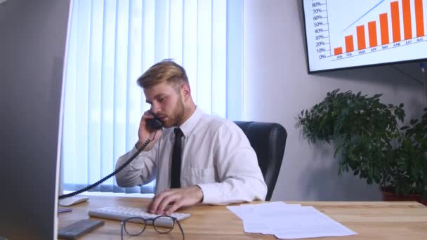 Jonge drukke zakenman praten op mobiele telefoon en kantoor telefoon zitten aan de tafel in office — Stockvideo