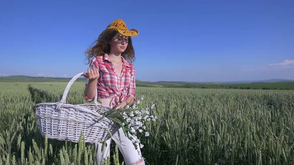 Молода жінка ходить з кошиком з квітами і пляшкою води пшеничне поле з блакитним небом на фоні. повільний рух — стокове фото