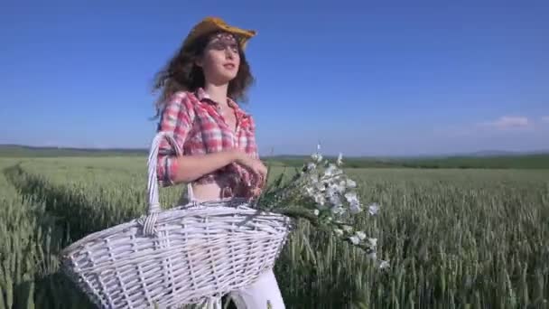 Młoda kobieta spaceru kosz z kwiatami i butelkę wody polu pszenicy z niebieski niebo na tle. Dziewczyna pić wodę z butelki. zwolnionym tempie — Wideo stockowe