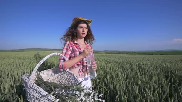 Eine junge Frau geht mit einem Korb mit Blumen und einer Flasche Wasser durch ein Weizenfeld mit blauem Himmel im Hintergrund. Mädchen trinken Wasser aus der Flasche. Zeitlupe — Stockvideo