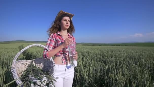 Молодая женщина идет с корзиной с цветами и бутылкой воды пшеничное поле с голубым небом на заднем плане. Девушка пьет воду из бутылки. замедленное движение — стоковое видео