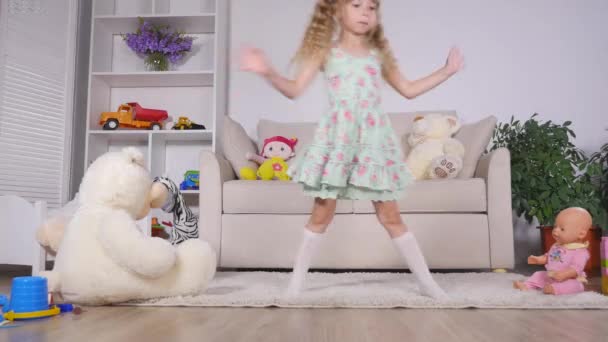 Ευτυχισμένος ξανθό κορίτσι στο φόρεμα διασκεδάζουν χορεύοντας σε εσωτερικούς χώρους σε ένα ηλιόλουστο δωμάτιο στο σπίτι με το λευκό ή το νηπιαγωγείο — Αρχείο Βίντεο