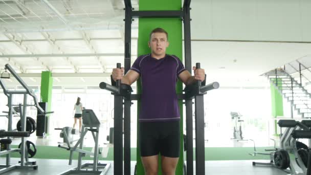 Inclinado hacia abajo de un joven barbudo con un torso desnudo haciendo levantamiento de piernas en una barra horizontal en un gimnasio — Vídeo de stock