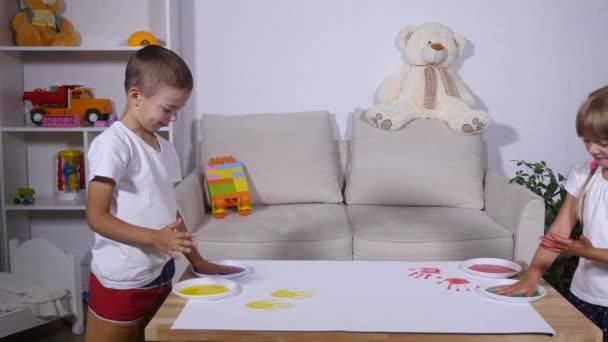 Un niño divertido lindo feliz pintando huellas de manos de color en la hoja blanca de papel — Vídeos de Stock