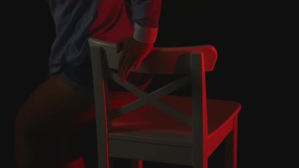 Ένα σέξι κορίτσι που κάθεται σε μια καρέκλα σε ένα σε σύντομο σορτς και ένα πουκάμισο σε μαύρο φόντο — Αρχείο Βίντεο