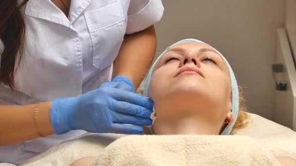 Una mujer bonita con los ojos cerrados, acostada en la mesa de los cosmetólogos, el dermatólogo usando esponjas de algodón para limpiar profesionalmente la cara del paciente. Cuidado facial, ser feliz, estar sano — Vídeos de Stock