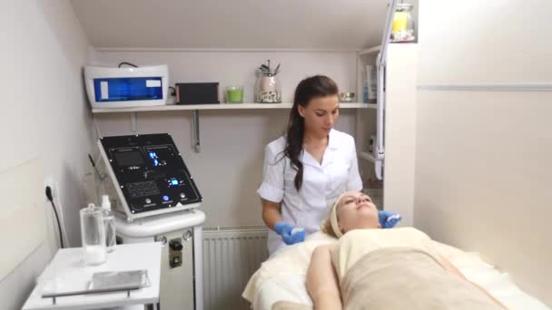 Una mujer bonita con los ojos cerrados, acostada en la mesa de los cosmetólogos, el dermatólogo usando esponjas de algodón para limpiar profesionalmente la cara del paciente. Cuidado facial, ser feliz, estar sano — Vídeo de stock