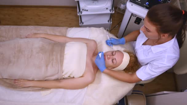 Jovem mulher bonita deitada na mesa clínica, o cosmetologista faz dela uma massagem de eletroestimulação facial. Procedimento de elevação, cosmetologia de hardware. Estilo de vida luxuoso, atividades modelo. Topo — Vídeo de Stock