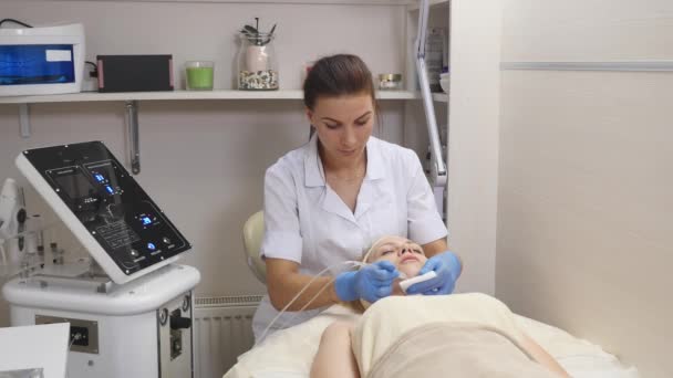 Giovane bella donna sdraiata sul tavolo clinico, la cosmetologa le fa un massaggio di elettrostimolazione facciale. Procedura di sollevamento, cosmetologia hardware. Stile di vita di lusso, attività modello — Video Stock