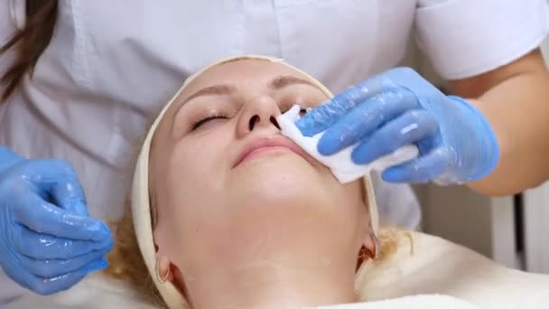 A pretty woman with closed eyes, lying on the cosmetologists table, the dermatologist using cotton sponges to professionally clean the face of the patient. Facial care, being happy, being healthy — Stock Video
