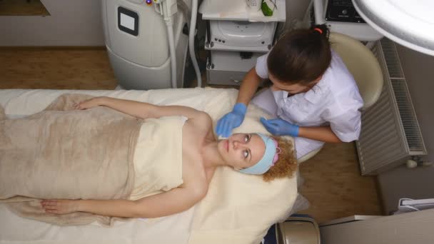A pretty woman with closed eyes, lying on the cosmetologists table, the dermatologist using cotton sponges to professionally clean the face of the patient. Facial care, being happy, being healthy. Top — Stock Video