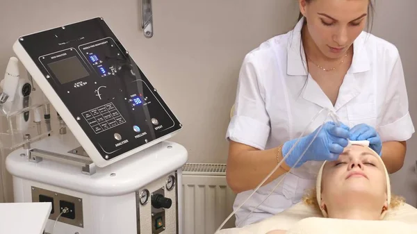 Joven hermosa mujer acostada en la mesa clínica, el cosmetólogo le hace un masaje de electro-estimulación facial. Procedimiento de elevación, cosmetología de hardware. Estilo de vida de lujo, actividades modelo — Foto de Stock