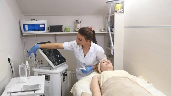 Joven hermosa mujer acostada en la mesa clínica, el cosmetólogo le hace un masaje de electro-estimulación facial. Procedimiento de elevación, cosmetología de hardware. Estilo de vida de lujo, actividades modelo — Foto de Stock