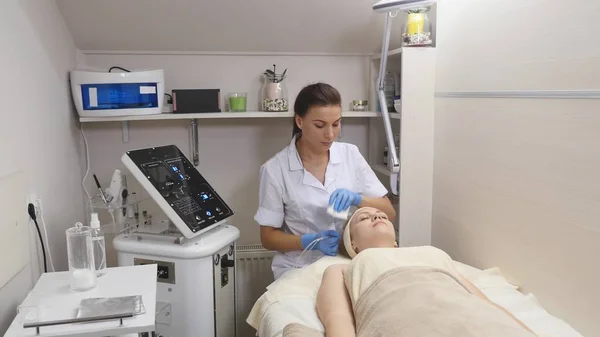 Joven hermosa mujer acostada en la mesa clínica, el cosmetólogo le hace un masaje de electro-estimulación facial. Procedimiento de elevación, cosmetología de hardware. Estilo de vida de lujo, actividades modelo — Foto de Stock