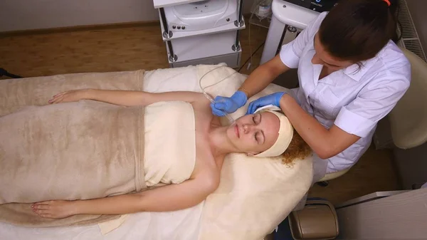 Joven hermosa mujer acostada en la mesa clínica, el cosmetólogo le hace un masaje de electro-estimulación facial. Procedimiento de elevación, cosmetología de hardware. Estilo de vida de lujo, actividades modelo. Parte superior — Foto de Stock