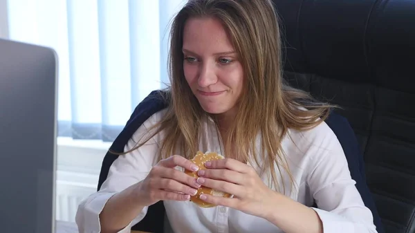 Młoda kobieta siedzi w biurze i jedzenia hamburgera — Zdjęcie stockowe