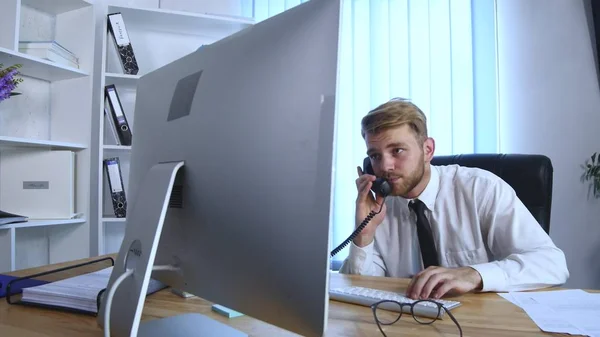 Молодой занятой бизнесмен разговаривает по мобильному телефону и офисному телефону и делает заметки в блокноте, сидя за столом в офисе — стоковое фото