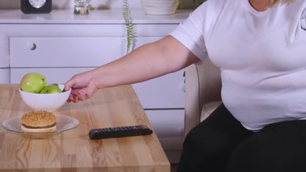 Толстая женщина ест бургер и смотрит телевизор — стоковое видео