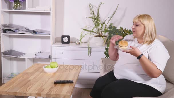 Mulher gorda comer um hambúrguer, assistindo TV e ri — Vídeo de Stock