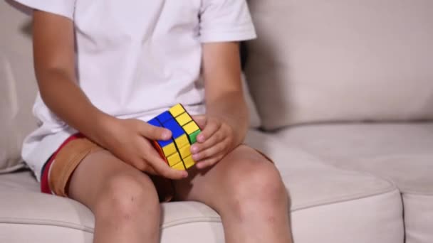 Il ragazzo è seduto sul divano e raccoglie un cubo di rubics — Video Stock