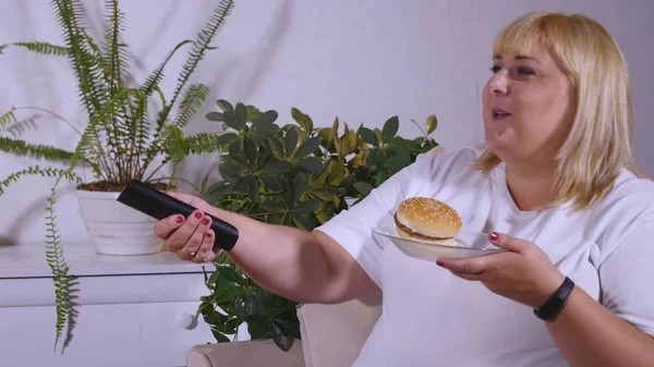 Gruba kobieta jedzenie hamburgera, oglądanie Tv i laughes — Zdjęcie stockowe