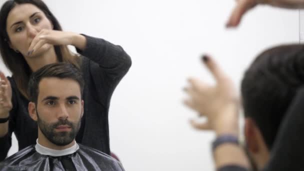 Cheerful skillful barber making a haircut with scissors to a young bearded man — Stock Video