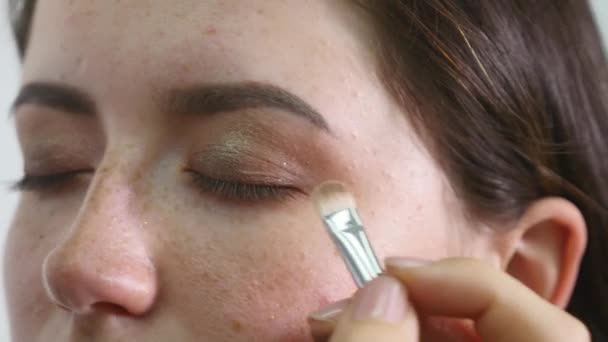 El primer plano del rostro del fotomodel joven profesional femenino durante el proceso del maquillaje en el salón de belleza. Visagiste joven está aplicando sombra de ojos en el párpado con un cepillo especial — Vídeo de stock