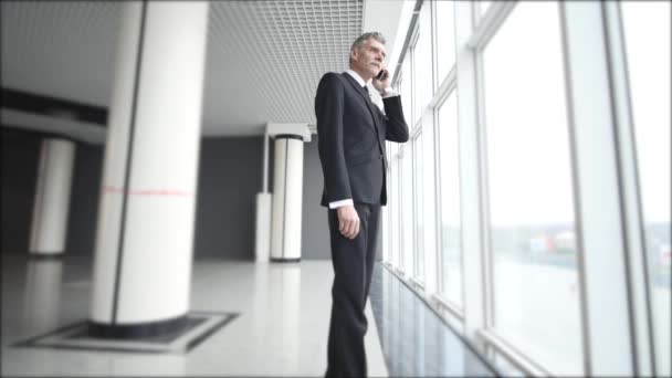 El viejo hombre de negocios habla por teléfono cerca de las ventanas panorámicas. Hombre hablando en un teléfono celular con una mirada seria de cara — Vídeo de stock
