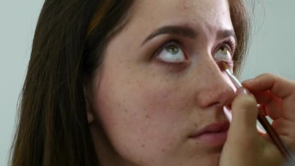 Close-up de uma cara de fotomodelo feminino profissional jovem durante o processo de maquilagem no salão de beleza. Young visagiste está aplicando sombra na pálpebra usando escova especial — Vídeo de Stock