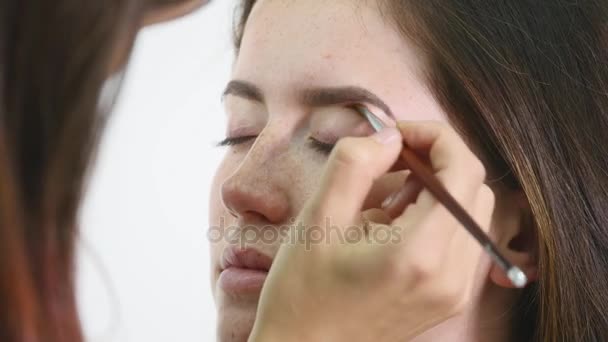 Närbild på ett ansikte av unga professionella kvinnliga photomodel under make-up på skönhetssalong. Unga visagiste tillämpar ögonskugga på ögonlocket med hjälp av speciell borste — Stockvideo