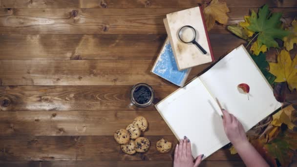 Herbstliches Konzept von oben. Bücher, Ahornblätter, Tee auf dem alten Holztisch. Frau schreibt Notizen ins Notizbuch — Stockvideo