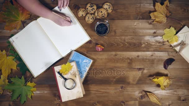 Conceito de outono vista superior. Livros, folhas de bordo, chá na velha mesa de madeira. Mulher escrevendo notas no caderno — Vídeo de Stock