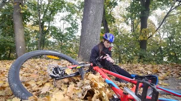 Une jeune femme s'assoit dans un parc après être tombée d'un vélo, calme la douleur dans son genou, est une balade dangereuse à vélo dans le parc un jour d'automne — Video