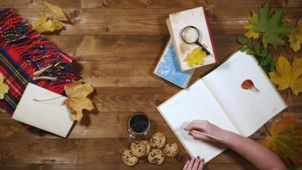Otoño concepto vista superior. Libros, hojas de arce, té en la vieja mesa de madera. Mujer escribiendo notas en el cuaderno — Vídeo de stock