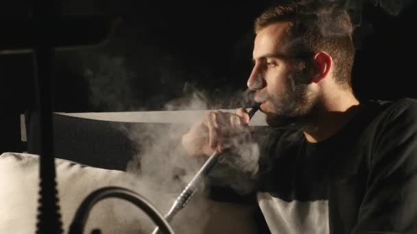 Молодой человек курит кальян в одиночестве на черном фоне — стоковое видео