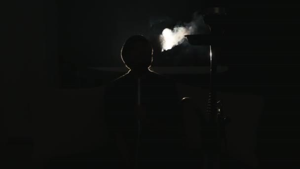 Young man smokes hookah and lets out smoke rings in a dark room close up — Stock Video