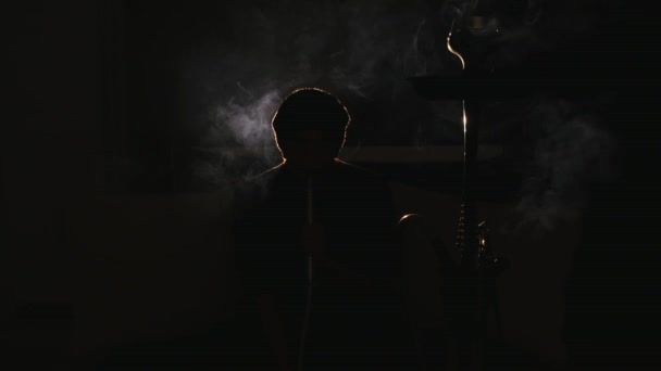 Jonge man rookt waterpijp en laat uit rook ringen in een donkere kamer close-up — Stockvideo