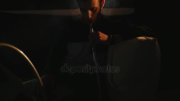Jeune homme fume narguilé et laisse sortir des anneaux de fumée dans une pièce sombre gros plan — Video