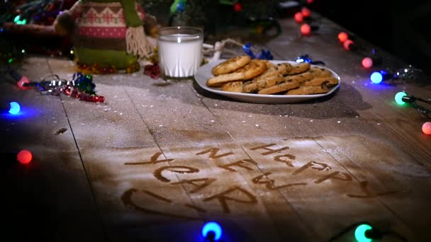 Fából készült barna asztal díszített koszorúkat és karácsonyi cucc. Egy pohár tej és a cookie-kat a karácsonyi asztal egy tányér — Stock videók