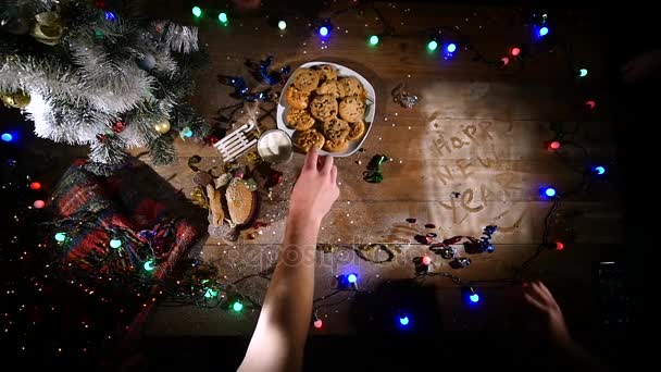 Houten bruine tafel versierd met Kerstmis spullen en slingers. handen neemt cookies van een plaat — Stockvideo